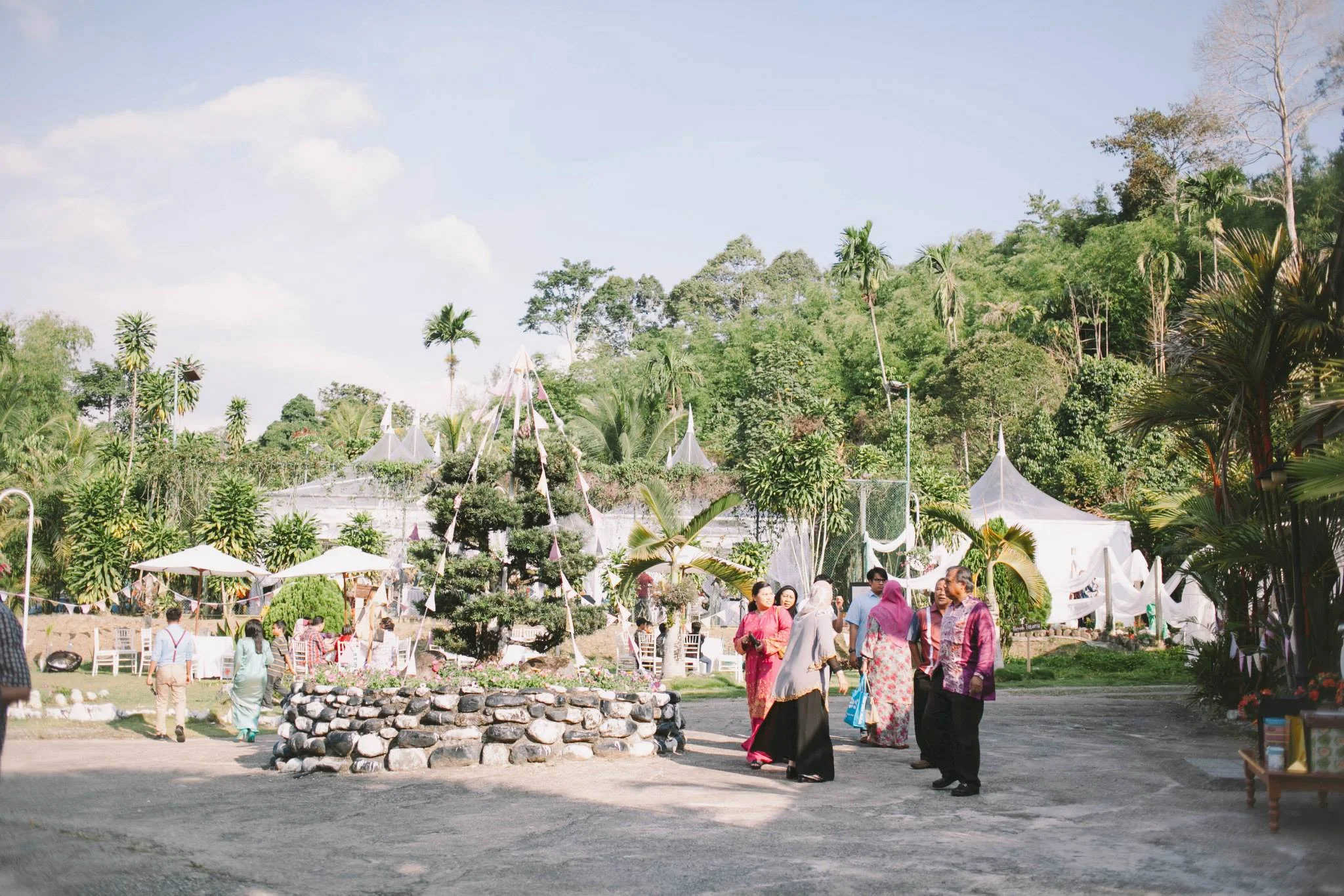 Guests in baju kurung and batik gathering on the grounds for an event, with white canopy tents